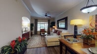 This single-floor residence offers a cohesive and elegantly designed interior defined by warm tones, rich wood accents, and thoughtful architectural details. The open-concept living and dining area serves as the heart of the home, featuring hardwood-style flooring, crown molding, and a ceiling fan overhead. The living room is anchored by a plush sectional sofa atop a floral area rug, complemented by a large decorative mirror and arched wall niche. Large windows dressed with drape panels invite natural light throughout. The dining area transitions seamlessly via an arched passageway, with a pendant light fixture above a wood dining table, and a console table and oval mirror adorning the entry hall. The kitchen is a standout, showcasing rich dark cherry cabinetry, granite countertops, stainless steel appliances including a French door refrigerator and built-in microwave, and a curved breakfast bar with leather-topped stools. Two bedrooms offer plush carpeting, ceiling fans, and arched windows. The primary bedroom features dark wood furniture and warm earth-toned bedding, while the second bedroom mirrors the same comfortable aesthetic. The bathroom includes a soaking tub with elegantly tiled travertine surround, completing this refined and functional home.