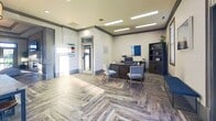 This 1000 square foot single-story office space in Glenmoore, PA, combines functionality with modern aesthetics. The interior showcases a striking herringbone pattern wood-look flooring throughout, creating a cohesive and sophisticated foundation. Recessed lighting and statement pendant fixtures provide ample illumination, complemented by natural light from large windows and glass doors.

The space is thoughtfully divided into distinct areas. A reception area features a dark wood desk, blue accent chairs, and built-in storage. The lounge space is anchored by a plush navy sofa and a large tufted ottoman, with a fireplace and mounted TV adding warmth and entertainment options. A central workspace area includes a table with blue chairs, ideal for collaborative work or meetings.

Architectural details such as tray ceilings with crown molding, accent walls with wood paneling, and strategically placed mirrors enhance the sense of space and style. The color palette primarily consists of neutral tones with pops of blue, creating a calming yet professional atmosphere. Despite its compact size, the layout maximizes functionality while maintaining an open, airy feel, making it an ideal setting for a modern business environment.