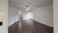This single-story space is designed with a clean, neutral palette and a practical layout visible across its main living areas, bedrooms, kitchen, and bathroom. The entry area features light tile flooring that transitions into rich, dark hardwood in the main living space, creating a clear visual distinction between zones. Crown molding runs along the ceilings throughout, adding a refined architectural detail, while ceiling fans with integrated lighting fixtures are present in multiple rooms, contributing both function and a consistent design element.

The living area is generously proportioned, offering an open feel enhanced by vertical blinds that filter natural light from the windows. A carpeted bedroom provides a comfortable, quiet retreat and includes a wall-mounted air conditioning unit and a full-width mirrored sliding closet, maximizing both storage and the sense of space. An additional carpeted room, also with a ceiling fan and window, offers similar comfort.

The kitchen is equipped with white cabinetry featuring brushed nickel hardware, dark granite countertops, a gas range, an over-the-range microwave, and ample upper and lower cabinet storage. The bathroom includes tile flooring and is finished in neutral tones with chrome accessories. Overall, the space presents a tidy, functional environment with consistent finishes throughout.