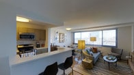 This well-designed single-floor residence showcases a seamless open-concept layout that integrates the kitchen, dining, and living areas into one cohesive and functional space. The kitchen is outfitted with modern cabinetry in warm wood tones, stainless steel appliances including a microwave and oven, and a generous white quartz peninsula with bar seating — ideal for casual dining or entertaining. Adjacent to the kitchen, a dedicated dining area provides a comfortable setting for meals. The living room is the highlight of the space, anchored by a large floor-to-ceiling window that floods the interior with abundant natural light and offers sweeping elevated views of the surrounding cityscape. The room comfortably accommodates a contemporary sofa, accent chairs, and stylish lighting fixtures that contribute to the warm, inviting ambiance. Light hardwood flooring flows throughout, complementing the neutral color palette of the walls and ceilings. The overall aesthetic is clean and modern, blending functional design with thoughtful décor accents including framed wall art and patterned area rugs. This space is well-suited for those seeking a stylish urban living environment with an emphasis on openness, light, and city connectivity.