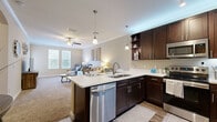 Located in the Highwoods Preserve area of Tampa, FL, this 1-bedroom, 1-bathroom residence offers a thoughtfully designed single-floor layout that maximizes comfort and functionality. The open-concept kitchen and living area serves as the heart of the home, where rich dark-wood cabinetry, stainless steel appliances, and bright white countertops create a striking visual contrast. A peninsula with bar seating and pendant lighting adds both style and practicality, while wood-look flooring grounds the kitchen space. Crown molding and a ceiling fan enhance the spacious living room, which is bathed in natural light from multiple windows and features plush carpet underfoot. The bedroom is a serene retreat with soft carpet, dual windows with blinds for privacy, and ample space for a full furniture arrangement. The bathroom is well-equipped with a dark-wood vanity, white countertop, a shower-tub combo, and wood-look flooring. A dedicated in-unit laundry room with side-by-side washer and dryer adds everyday convenience. Throughout the home, warm neutral tones, crown molding, and consistent finishes contribute to a cohesive and inviting ambiance.
