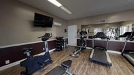 Located at 1503 E Park Ave in Valdosta, Georgia, this fitness center offers a well-equipped, single-floor exercise facility designed to support a range of workout routines. The room features wood-look flooring throughout, paired with a two-tone wall treatment — warm taupe on the upper half and deep burgundy on the lower — giving the space a clean, purposeful aesthetic.

The facility is outfitted with a comprehensive selection of cardio equipment, including Spirit-brand treadmills, an elliptical trainer, and stationary bikes. A multi-functional cable machine and a weight bench provide options for strength training, while a standing dumbbell rack with multiple weight increments supports free-weight exercises. A wall-mounted flat-screen TV offers entertainment during workouts, and a mini-split HVAC unit ensures climate control year-round.

Natural light enters through multiple windows fitted with slatted blinds, brightening the space alongside overhead fluorescent lighting. A large wall mirror on one side visually expands the room and allows users to monitor their form. Fitness center rules are posted near the entrance, promoting a safe and orderly environment. The single-door entry provides straightforward access, making this a practical and functional fitness amenity.