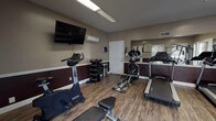 Located at 1503 E Park Ave in Valdosta, Georgia, this fitness center offers a well-equipped, single-floor exercise facility designed to support a range of workout routines. The room features wood-look flooring throughout, paired with a two-tone wall treatment — warm taupe on the upper half and deep burgundy on the lower — giving the space a clean, purposeful aesthetic.

The facility is outfitted with a comprehensive selection of cardio equipment, including Spirit-brand treadmills, an elliptical trainer, and stationary bikes. A multi-functional cable machine and a weight bench provide options for strength training, while a standing dumbbell rack with multiple weight increments supports free-weight exercises. A wall-mounted flat-screen TV offers entertainment during workouts, and a mini-split HVAC unit ensures climate control year-round.

Natural light enters through multiple windows fitted with slatted blinds, brightening the space alongside overhead fluorescent lighting. A large wall mirror on one side visually expands the room and allows users to monitor their form. Fitness center rules are posted near the entrance, promoting a safe and orderly environment. The single-door entry provides straightforward access, making this a practical and functional fitness amenity.