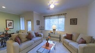 This single-floor residence offers a warm and comfortable living environment defined by its light, neutral color palette and thoughtful layout. The living room serves as the heart of the home, featuring plush seating arranged around a glass-top coffee table, creating an inviting space for relaxation and conversation. Large windows dressed with elegant curtain valances allow generous natural light to fill the room, offering views of the surrounding greenery outdoors. A ceiling fan with a central light fixture provides both comfort and illumination, while the soft carpeted flooring adds a sense of warmth underfoot. Decorative touches, including accent pillows in earthy tones, a ceramic flower vase, and framed artwork on the walls, contribute to the space's cozy and personalized character. A glass-and-metal console table beneath the main window provides a functional yet stylish accent, complemented by a classic table lamp that adds a soft glow to the room. Vertical blinds on side windows offer additional light control and privacy. The overall ambiance of the space is relaxed and homey, making it well-suited for comfortable everyday living.
