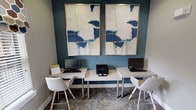 Located at 8630 Spring Garden Drive in Fort Worth, Texas, this single-floor business center offers a functional and thoughtfully designed workspace. The room centers around a pair of sleek white workstations, each equipped with a desktop computer and ergonomic modern chair, with a shared printer positioned conveniently between them. The workstation area is anchored by a deep teal accent wall that provides a bold visual backdrop, adorned with two large-format abstract art pieces in gold frames — featuring navy, teal, and white tones that lend the space a contemporary, polished character. The remaining walls are finished in a soft neutral gray, creating a calm and focused atmosphere. Natural light filters into the space through windows dressed with white horizontal blinds and decorative hexagonal-patterned fabric valances in navy, brown, and cream tones, adding warmth and texture. The flooring features a subtly patterned carpet that complements the overall color palette. A glass-paneled wooden door with an automatic closer provides access to the exterior, offering views of landscaped grounds and a gated outdoor area. The space is well-suited for productivity, balancing aesthetic appeal with practical amenities in a clean, organized environment.