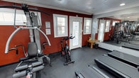This dedicated fitness center is thoughtfully designed to accommodate a full range of exercise needs within a single, efficiently organized space. The room features a bold two-tone color scheme, with deep red accent walls contrasting against neutral gray walls, crown molding, and a dark rubberized floor that provides durability and traction underfoot. Recessed lighting illuminates the space evenly, while multiple windows along the red accent wall allow natural light to filter in, creating an energizing atmosphere. A large floor-to-ceiling mirror spans one full wall, visually expanding the room and allowing users to monitor their form during workouts. The cardio section is well-stocked with multiple treadmills from Life Fitness and NordicTrack, an elliptical trainer, and a spin bike, offering a variety of aerobic training options. A multi-function cable and weight machine serves as the primary strength training station, complemented by additional resistance equipment. A wall-mounted flat-screen television provides entertainment during workouts. A glass-paneled interior door connects the fitness center to an adjacent common area, and an exterior door offers convenient access to the outdoors. A small wooden side table and waste bin round out the practical amenities of this clean and functional facility.