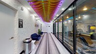 This fitness center is a thoughtfully designed facility that combines striking aesthetics with comprehensive workout functionality. The entrance corridor immediately captures attention with its dramatic wave-form wooden ceiling installation, backlit with vibrant multicolored LED lighting that transitions through purple, pink, and green hues. The hallway is lined with a striped runner rug, textured accent walls, and floor-to-ceiling glass panels that offer a clear sightline into the main workout floor, creating a sense of openness and anticipation.

Access to the facility is provided via an elevator lobby, which shares the same artistic ceiling design, maintaining visual continuity throughout. The main fitness floor is expansive, featuring dark rubber flooring and a dark dropped ceiling with recessed and panel lighting. Walls are painted in a cool teal-gray tone, contributing to a focused and energizing atmosphere.

The cardio zone is well-stocked with treadmills, stationary bikes, elliptical trainers, and rowing machines, positioned along large windows that flood the space with natural light and provide views of the surroundings. The strength training area includes a variety of selectorized cable machines, weight benches, a full dumbbell rack, kettlebells, and additional functional training equipment. A wall-mounted flat-screen TV is also present. Changing room doors are accessible from within the fitness floor, completing this full-service facility.