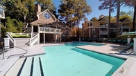 Located at 6713 Six Forks Rd in Raleigh, NC, this outdoor amenity area centers around a generously sized swimming pool that serves as the focal point of the space. The pool is flanked by a wide concrete pool deck lined with teal lounge chairs and shaded by market umbrellas, offering ample space for sunbathing and relaxation. A covered pavilion area at the pool's edge provides a shaded retreat with seating including bistro tables and lounge chairs, ideal for gathering out of direct sunlight. Adjacent to the pool deck is a raised upper-level deck, finished with white railings and furnished with bright green cushioned seating, offering elevated views of the pool and surrounding landscape. The clubhouse building features traditional architecture with a pitched gable roof, a prominent dormer window, and a chimney, clad in warm taupe lap siding. The entire pool area is enclosed by a black iron fence for safety and privacy. Tall mature pine and hardwood trees frame the perimeter of the space, creating a serene, nature-filled backdrop. The grounds are well-maintained, and the overall ambiance is relaxed and resort-like, blending functional amenities with an inviting outdoor atmosphere.