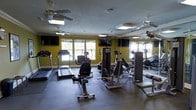 This fitness center is designed to support a full range of workout routines in a comfortable, functional environment. The space is outfitted with an array of cardio equipment, including treadmills, elliptical trainers, and a rowing machine, alongside dedicated strength training stations to accommodate varied fitness goals. The flooring features durable rubber gym matting, providing safety and stability throughout the workout area. Large windows line the walls, inviting generous amounts of natural light and creating an open, airy atmosphere. Bold yellow accent walls add energy to the space, complementing the modern, purpose-driven aesthetic. Multiple ceiling fans are distributed across the room to ensure consistent airflow and comfort during exercise. Wall-mounted flat-screen televisions are positioned throughout, allowing users to stay entertained or informed while training. A floor-to-ceiling mirror along one wall visually expands the space and allows for form checks during workouts. Crown molding along the upper walls adds a refined architectural touch, elevating the overall design of the room. The single-level layout ensures seamless accessibility, making this fitness center a practical and well-appointed facility for daily use.