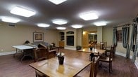 This single-floor space offers a warm and functional environment designed for communal gathering and dining. The open layout seamlessly connects a lounge area with a dining space, both finished with rich hardwood flooring that adds a sense of warmth throughout. Multiple large flush-mount ceiling light panels provide bright, even illumination across the entire area, ensuring a well-lit and welcoming atmosphere at all times.

The lounge area features comfortable seating, including wicker-style armchairs and a sofa, arranged around a central focal wall. This wall showcases a built-in entertainment unit with a flat-screen television, flanked by decorative shelving niches ideal for displaying plants and ornamental pieces. A cabinet unit with storage drawers sits alongside, adding practical functionality. A framed artwork piece adorns the adjacent wall, contributing to the homey aesthetic.

The dining area accommodates multiple wooden tables and chairs, arranged to support both individual and group seating. Potted floral arrangements are placed throughout, adding natural color and vibrancy to the space. A doorway in the background leads to an additional room, visible as a secondary lounge or library area with warm amber lighting and shelving. The overall ambiance is inviting, practical, and thoughtfully arranged for daily communal use.