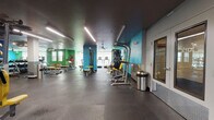 This fitness center is a single-floor facility designed to support a comprehensive workout experience across several distinct zones. The space features durable rubber flooring throughout and a dark ceiling fitted with linear LED lighting, creating a modern, energetic atmosphere.

The cardio area is positioned along a windowed wall, housing a row of Matrix treadmills and elliptical trainers alongside a stationary bike, including a Peloton. Large windows allow natural light to fill this zone and offer outdoor views, enhancing the environment during exercise. Full-length mirrors run along one wall, visually expanding the space.

The strength training area includes a dumbbell rack with a flat bench, multiple selectorized weight machines with yellow upholstery, a cable tower, and a leg extension machine. A functional training station with medicine balls, resistance bands, and stability balls of varying sizes is organized on a dedicated rack along one wall.

Vibrant teal and green accent walls energize the space and define different workout zones. Amenities include a water fountain station, restroom access, a sanitation dispenser, and waste bins placed throughout. Ceiling fans provide additional ventilation in select areas. The layout offers ample open floor space, ensuring comfortable movement between equipment stations.