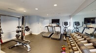 Located at 1834 Jackson Bluff Rd in Tallahassee, FL, this single-floor fitness facility is thoughtfully designed to support a full range of exercise disciplines. The space is divided into distinct functional zones, each finished with light blue walls, recessed lighting, and clean white trim throughout.

The primary workout area features rubber flooring suited for heavy training, housing a Smith machine with a weight bench, two treadmills with individual display screens, a wall-mounted flat-screen TV, and a cable machine. A full-length mirror spans one wall, amplifying the sense of space and aiding form during exercise. A rack of hex dumbbells in graduated sizes and a collection of kettlebells provide comprehensive free weight options, while a upright bike rounds out the cardio selection.

A separate studio room offers a distinct environment for yoga, cycling, and group fitness. This area is finished with warm, reclaimed-style hardwood floors and features large wall mirrors, a wall-mounted TV, spin bikes, and open floor space ideal for mat-based workouts or stretching. Stability balls and additional accessories are neatly stored on shelving within this room.

Connecting the two zones is a transitional area with frosted glass-panel doors, maintaining visual openness while defining each space. Natural light filters in through slatted blinds, and safety equipment including a fire extinguisher and emergency exit signage are clearly visible throughout.