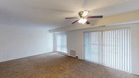 This single-floor space presents a clean, functional living area designed with comfort and practicality in mind. The room is finished with wall-to-wall beige carpeting, creating a warm and inviting atmosphere underfoot. A central ceiling fan with an integrated light fixture serves as the focal point overhead, providing both ambient lighting and air circulation throughout the space.

A defining feature of the room is the expansive wall of vertical blinds, which spans two large openings — a wide window and what appears to be a sliding glass door — allowing abundant natural light to filter into the interior while offering privacy control. The sliding glass door provides convenient access to an outdoor area, visible through the blinds.

The walls are finished in a neutral white tone, complementing the light-colored ceiling and contributing to an open, airy feel. A structural beam detail runs along the upper wall, adding a subtle architectural element. A floor-level air vent is positioned near the base of the window wall, ensuring efficient climate control throughout the space. Overall, the room offers a straightforward, versatile layout well-suited for a variety of furnishing arrangements.