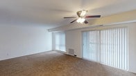This single-floor space presents a clean, functional living area designed with comfort and practicality in mind. The room is finished with wall-to-wall beige carpeting, creating a warm and inviting atmosphere underfoot. A central ceiling fan with an integrated light fixture serves as the focal point overhead, providing both ambient lighting and air circulation throughout the space.

A defining feature of the room is the expansive wall of vertical blinds, which spans two large openings — a wide window and what appears to be a sliding glass door — allowing abundant natural light to filter into the interior while offering privacy control. The sliding glass door provides convenient access to an outdoor area, visible through the blinds.

The walls are finished in a neutral white tone, complementing the light-colored ceiling and contributing to an open, airy feel. A structural beam detail runs along the upper wall, adding a subtle architectural element. A floor-level air vent is positioned near the base of the window wall, ensuring efficient climate control throughout the space. Overall, the room offers a straightforward, versatile layout well-suited for a variety of furnishing arrangements.