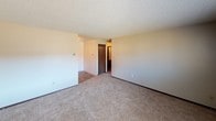 This single-floor residential unit offers a practical and straightforward layout designed for comfortable everyday living. The living area is generously sized, featuring soft wall-to-wall carpeting, neutral beige walls, and a textured ceiling. A wall-mounted air conditioning unit and baseboard heating provide climate control throughout the season, while a broad window invites natural light and outdoor views.

The kitchen is efficiently arranged with dark wood cabinetry lining both sides of the space, offering ample storage. White laminate countertops, a coil-burner electric range, a range hood, and a stainless steel sink complete the functional setup. Durable tile flooring ties the kitchen together and provides easy maintenance.

The bedrooms are well-proportioned, each finished with plush carpeting, neutral walls, and textured ceilings. Baseboard heating runs along the exterior walls, and windows bring in natural daylight. Each bedroom also features closet access and dark wood-trimmed doors consistent with the unit's overall aesthetic.

The bathroom is clean and functional, featuring a full soaking tub with a tiled surround in warm neutral tones, a standard toilet, and a towel bar. The overall unit presents a consistent, no-frills design with warm neutral tones throughout, making it a practical and move-in ready living space.