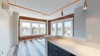 This single-floor urban unit masterfully combines industrial character with modern design. The open living and kitchen area is a standout, showcasing exposed red brick walls on multiple sides and a generous bank of white-framed windows that frame panoramic rooftop views of the surrounding cityscape. Sunlight pours in throughout the day, highlighting the light-toned luxury vinyl plank flooring and the bold orange conduit that traces the ceiling — a deliberate industrial design element. A wall-mounted mini-split HVAC unit ensures efficient climate control.

The kitchen is thoughtfully appointed with two-toned cabinetry — dark navy lower cabinets paired with light grey uppers — complemented by crisp white quartz countertops, a matte black faucet, stainless steel appliances including a range, microwave, and refrigerator, and a single-basin undermount sink.

The bedroom offers a quieter retreat, featuring soft carpeting underfoot, an exposed brick accent wall, and a bright double-pane window with a lush tree-lined view. The bathroom is clean and functional, finished with large-format tile, a natural light window with frosted glass, and matte black hardware accents throughout. The overall ambiance is a refined balance of raw, urban texture and polished contemporary living.