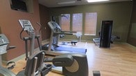 This fitness center offers a practical and well-organized space designed to support a comprehensive workout routine. The room features warm hardwood-style flooring that contrasts with the earthy, deep green-toned walls, creating a focused and energizing atmosphere. Natural light filters in through a row of windows fitted with horizontal blinds, providing a bright yet comfortable environment for exercise.

The facility is outfitted with a solid selection of cardio equipment, including Life Fitness elliptical trainers and treadmills positioned to take advantage of the natural light from the windows. A rowing machine is also available, adding variety to the available cardio options. A multi-station cable machine is visible toward the back of the room, offering strength training capabilities alongside the cardio equipment. A flat-screen television is mounted in the corner, allowing users to stay entertained during their workouts.

The layout of the space is efficient and thoughtfully arranged, ensuring adequate room to move between machines. Mirrored wall panels add depth to the room and allow users to monitor their form during exercise. The overall ambiance is clean, functional, and well-maintained, making it a capable facility suited to a wide range of fitness goals and routines.
