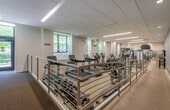 This fitness center offers a comprehensive workout environment designed to accommodate a wide range of exercise needs. The space is laid out across a single floor and is organized into distinct functional zones, allowing users to move seamlessly between cardio, strength training, and free weight activities.

The cardio section is well-stocked with Life Fitness treadmills, elliptical trainers, and stationary bikes, all positioned to take advantage of natural light streaming in through large windows that offer views of surrounding greenery. The strength training area features a variety of cable machines and multi-functional equipment, including a Synrgy360 functional training station, providing versatile options for resistance-based workouts. An adjustable weight bench and free weights round out the strength offerings.

The facility is finished with durable speckled rubber flooring throughout, well-suited for high-traffic use. Full-length mirrors line portions of the walls, enhancing the sense of space and allowing users to monitor their form. Overhead, a suspended ceiling with linear LED lighting ensures bright, even illumination across the entire floor.

Additional amenities include a wall-mounted water fountain, a yoga mat, a stability ball, and informational postings. The muted blue-gray accent walls and neutral tones contribute to a clean, focused atmosphere conducive to productive workouts.