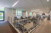 This fitness center offers a comprehensive workout environment designed to accommodate a wide range of exercise needs. The space is laid out across a single floor and is organized into distinct functional zones, allowing users to move seamlessly between cardio, strength training, and free weight activities.

The cardio section is well-stocked with Life Fitness treadmills, elliptical trainers, and stationary bikes, all positioned to take advantage of natural light streaming in through large windows that offer views of surrounding greenery. The strength training area features a variety of cable machines and multi-functional equipment, including a Synrgy360 functional training station, providing versatile options for resistance-based workouts. An adjustable weight bench and free weights round out the strength offerings.

The facility is finished with durable speckled rubber flooring throughout, well-suited for high-traffic use. Full-length mirrors line portions of the walls, enhancing the sense of space and allowing users to monitor their form. Overhead, a suspended ceiling with linear LED lighting ensures bright, even illumination across the entire floor.

Additional amenities include a wall-mounted water fountain, a yoga mat, a stability ball, and informational postings. The muted blue-gray accent walls and neutral tones contribute to a clean, focused atmosphere conducive to productive workouts.