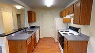 This single-floor residential unit offers a practical and comfortable layout designed for easy everyday living. The kitchen is a functional galley-style space outfitted with warm oak cabinetry, gray laminate countertops, a double basin sink, and white appliances including a coil-burner electric range and dishwasher. Durable hardwood-style flooring runs throughout the kitchen, which connects seamlessly to the open living and dining area. The living area is generously sized with plush wall-to-wall carpeting and benefits from multiple windows that invite natural light and offer views of the surrounding greenery. A pendant light fixture above the kitchen peninsula serves as a subtle design accent connecting the two spaces. The bedrooms are carpeted and bright, with one featuring a large window framing an outdoor view of mature trees. The primary bedroom stands out with direct access to a well-appointed en-suite bathroom, complete with warm wood vanity cabinetry and a lighted mirror, as well as a spacious walk-in closet. Throughout the unit, neutral wall tones and clean lines create a calm, move-in-ready atmosphere suited to a variety of furnishing styles.