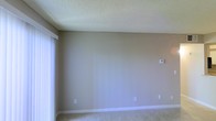 This single-story space presents a clean, move-in-ready interior with a straightforward and functional design. The room visible in the images features neutral beige walls that create a calm, versatile backdrop suitable for a variety of furnishing styles. Soft carpet flooring runs throughout, adding a sense of warmth and comfort underfoot.

The ceiling features a classic popcorn texture, a common finish that adds subtle dimension overhead. Natural light enters generously through a large window fitted with a white roller blind, brightening the space and lending an open, airy quality to the interior. White baseboards provide a crisp contrast against the neutral walls, contributing to a tidy, well-maintained appearance.

Practical conveniences are in place, including multiple electrical outlets positioned along the walls and a wall-mounted thermostat, allowing for straightforward climate control. The room's clean lines and uncluttered layout offer a blank canvas that can be easily adapted to suit personal preferences. Overall, the space delivers a simple, functional, and comfortable environment with a neutral aesthetic.