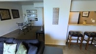 This single-floor space offers an open-concept layout that seamlessly connects the living and dining areas, creating a cohesive and functional environment. A structural column serves as a natural divider between zones while maintaining visual openness throughout the interior.

The living area features carpeted flooring and is furnished with a dark leather sofa accented by light-colored cushions. A decorative wine varietal art print — listing names such as Pinot Noir, Bordeaux, Merlot, and Malbec — is mounted on the column, adding a stylish, thematic touch to the space.

Beyond the column, the dining area accommodates a bistro-style table and chairs beneath a collection of framed wall art, with a floor lamp providing warm ambient lighting. A small side table with a decorative accent piece bridges the two zones.

To the right, a raised counter with a bar-height stool suggests a kitchen pass-through or bar area, enhancing the entertainment-friendly layout. The space benefits from neutral wall tones and carpeted floors that lend a warm, relaxed ambiance suited to both everyday living and casual hosting.