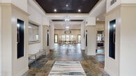 This single-story community clubhouse is thoughtfully designed to offer a range of amenities within a polished, welcoming environment. The grand lobby sets the tone upon entry, featuring multicolored slate tile flooring, a dramatic tray ceiling finished in deep charcoal, and a cascading chandelier that anchors the space. Structural columns with decorative trim frame sightlines toward a bright lounge area at the rear, furnished with seating and bathed in natural light from multiple windows and glass French doors that open to the outdoors.

Adjacent to the entry, a seating alcove with modern benches and wall sconces provides a refined waiting area, while a corridor with continued slate tile flooring leads to the facility's additional amenities. The conference room offers a wood-toned table surrounded by upholstered chairs and a wall-mounted display, creating a clean, functional setting for meetings.

The fitness center is generously proportioned, housing treadmills, ellipticals, a cable machine, free weights, and a flat-screen TV. Large windows allow natural light and outdoor views throughout. A kitchenette with bold granite countertops, a stainless steel wall oven, and pendant lighting provides a practical and attractive space for gatherings.