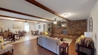 Located at 1651 Knollwood Dr in Mobile, Alabama, this single-story home offers a warm and inviting interior defined by thoughtful architectural details and a cohesive open layout. The living area is a standout space, anchored by a dramatic floor-to-ceiling stone fireplace with a wood mantle that serves as the room's focal point. Exposed wooden ceiling beams run the length of the room, adding rustic character and visual depth to the textured ceiling above. Rich hardwood flooring flows throughout the living area, complementing the natural tones of the stonework and beams. Ceiling fans provide practical comfort while enhancing the casual, lived-in atmosphere. The space transitions seamlessly into a dining area, where light tile flooring distinguishes the zone while maintaining an open, connected feel. Natural light filters in through windows near the dining space, brightening the interior and creating an airy ambiance. A doorway from the dining area leads to additional areas of the home, suggesting a well-connected floor plan. Together, the stone fireplace, exposed beams, and warm wood tones create a cohesive aesthetic that balances rustic charm with everyday comfort, making this home a welcoming retreat in Mobile, AL.