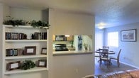 This single-floor residence offers a thoughtfully designed open-concept layout that promotes a seamless flow between living, dining, and kitchen areas. A defining architectural feature is the pass-through kitchen window, which creates a convenient visual and functional connection between the kitchen and the adjacent living space, making the layout feel open and interactive.

The kitchen is equipped with black appliances, including a gas range and an overhead microwave, set against warm wood-toned cabinetry and light countertops. The pass-through ledge doubles as a casual bar or serving counter, enhancing everyday functionality.

The living area is accented by a set of built-in white shelves, providing ample display and storage space for books, décor, and personal items. The textured popcorn ceiling is consistent throughout the space, adding a classic residential character.

Adjacent to the kitchen, the dining area benefits from generous natural light filtering through large windows dressed with light curtains, creating a bright and welcoming atmosphere. The dining space comfortably accommodates a table and chair set, making it well-suited for everyday meals or entertaining. Together, the rooms create a cohesive, functional, and comfortable living environment.