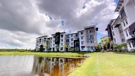 This contemporary residential building presents a striking exterior defined by a sophisticated contrast of white and dark facade panels, creating a bold and modern aesthetic. Rising several stories, the structure features multiple private balconies on each level, offering residents elevated views of the surrounding landscape. The building is nestled along the edge of a tranquil water feature, with lush green lawns sweeping down to the water's edge, enhancing the sense of a serene, resort-style setting. Tropical palm trees and mature landscaping are thoughtfully integrated throughout the grounds, reinforcing the warm, South Florida ambiance. Large windows are prominent across the facade, suggesting well-lit interiors that take advantage of the natural light and outdoor scenery. The architectural design blends clean, geometric lines with subtle traditional roofline details, striking a balance between modern and classic sensibilities. The waterfront positioning of the building adds a distinctive character to the property, with the reflective water views contributing to an overall atmosphere of calm and comfort. The single-floor unit within this building benefits from this carefully designed community setting, placing residents in close proximity to both natural surroundings and contemporary amenities.