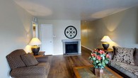 This single-story space offers a comfortable and well-appointed interior defined by rich, dark hardwood flooring that runs throughout the main living area, adding warmth and character to the overall design. The living room serves as the heart of the space, anchored by a tile-surround fireplace that creates a natural focal point and cozy atmosphere. Above the fireplace, a decorative wrought-iron medallion wall accent adds an artistic touch to the neutral-toned walls. The room is generously proportioned, providing ample space for seating arrangements and entertaining. Warm ambient lighting is provided by classic table lamps positioned on either side of the room, contributing to the inviting ambiance. A white interior door leads to an adjacent area, suggesting a well-connected floor plan. The neutral wall palette, ranging from soft white to warm beige, complements the earthy tones of the hardwood floors and wooden furniture pieces, including a tile-top coffee table that grounds the seating area. The single-floor layout ensures easy accessibility throughout, while the cohesive design elements create a space that feels both functional and homey.