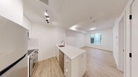 This efficiently designed one-bedroom, one-bathroom unit in Calgary, AB offers a thoughtfully laid-out single-floor plan with a clean, contemporary aesthetic. The open-concept living and kitchen area forms the heart of the unit, where light hardwood-style flooring runs throughout, complementing the crisp white walls and creating a bright, cohesive atmosphere. The kitchen is equipped with stainless steel appliances — including a refrigerator, stove, microwave, and dishwasher — paired with flat-panel white cabinetry and a white quartz peninsula that doubles as a workspace and casual dining area. Track lighting above the kitchen adds functional illumination, while a recessed ceiling detail adds subtle architectural interest. The living area benefits from a window overlooking a balcony railing, allowing natural light to filter in. The bedroom is a cozy, well-defined space with its own window view of an outdoor balcony area and light flooring consistent with the rest of the unit. The bathroom features a clean, all-white design with floor-to-ceiling white tile surrounding a soaker tub and shower combination fitted with chrome fixtures. Overall, the unit presents a modern, minimalist character with practical finishes suited for comfortable urban living.