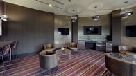 This single-floor lounge space showcases a refined, contemporary design with a distinctly upscale aesthetic. The room is anchored by a bold multi-toned striped carpet in shades of purple, burgundy, and grey, which runs throughout and provides a dynamic visual foundation. Dark, textured walls clad in metallic mosaic tile finishes add depth and a glamorous character to the interior.

The seating area features multiple curved, leather and woven lounge chairs arranged around transparent acrylic side tables, creating an inviting and flexible gathering environment. Overhead, polished chrome globe pendant lights are suspended at varying points, providing ambient illumination that complements the recessed ceiling lighting.

Two wall-mounted flat-screen televisions are integrated seamlessly into the dark paneling, serving different zones of the space. A built-in wet bar along one wall includes a curved stone countertop, an undermount sink, a microwave, and a compact refrigerator — all finished in dark cabinetry that echoes the room's color palette.

Large windows dressed with floor-length textured drapes allow natural light to filter in while offering exterior views. Along one wall, a narrow console bar table is accompanied by counter-height stools beneath a trio of framed abstract artworks, lit by individual pendant fixtures, adding a gallery-like touch to the overall ambiance.