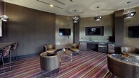 This single-floor lounge space showcases a refined, contemporary design with a distinctly upscale aesthetic. The room is anchored by a bold multi-toned striped carpet in shades of purple, burgundy, and grey, which runs throughout and provides a dynamic visual foundation. Dark, textured walls clad in metallic mosaic tile finishes add depth and a glamorous character to the interior.

The seating area features multiple curved, leather and woven lounge chairs arranged around transparent acrylic side tables, creating an inviting and flexible gathering environment. Overhead, polished chrome globe pendant lights are suspended at varying points, providing ambient illumination that complements the recessed ceiling lighting.

Two wall-mounted flat-screen televisions are integrated seamlessly into the dark paneling, serving different zones of the space. A built-in wet bar along one wall includes a curved stone countertop, an undermount sink, a microwave, and a compact refrigerator — all finished in dark cabinetry that echoes the room's color palette.

Large windows dressed with floor-length textured drapes allow natural light to filter in while offering exterior views. Along one wall, a narrow console bar table is accompanied by counter-height stools beneath a trio of framed abstract artworks, lit by individual pendant fixtures, adding a gallery-like touch to the overall ambiance.