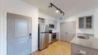 Located on W Kinderton Way in Bermuda Run, NC, this single-floor residence offers a thoughtfully designed open-concept layout with consistent hardwood flooring throughout. The kitchen is a functional highlight, outfitted with stainless steel appliances — including a full-size refrigerator, electric range, and built-in microwave — complemented by white subway tile backsplash, granite countertops, and gray shaker-style cabinetry. A decorative diamond-pattern wine rack accent adds character above the upper cabinets. Track lighting illuminates the kitchen and living areas, creating a bright, modern atmosphere. The kitchen opens seamlessly into the living space, which is generously sized and bathed in natural light from a set of French doors with a transom window that lead to an exterior balcony. Two bathrooms serve the unit: one features a tub-shower combination with a curved curtain rod, gray vanity cabinetry, and a granite countertop, while the second offers a walk-in shower stall with built-in shelving. A double-door closet in the kitchen area provides ample storage. The clean, neutral color palette of white walls and light wood floors gives the space a fresh, move-in-ready appeal throughout.