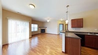 This single-floor space is designed with an open-concept layout that seamlessly connects the kitchen and living areas, creating a spacious and functional environment. The flooring throughout consists of warm, light-toned hardwood-style planks that lend a clean and inviting aesthetic to the space.

The kitchen area features rich, dark-stained cabinetry paired with a dark countertop surface that extends to form a breakfast bar or peninsula, offering casual seating potential and a natural division between the kitchen and the main living area. A pendant light fixture hangs above the counter, providing focused task lighting with a touch of style.

The living area is generously proportioned, anchored by a tiled fireplace that serves as a natural focal point. A ceiling fan with integrated lighting is centrally positioned overhead, promoting airflow and supplementary illumination. Vertical blinds dress a large window, allowing for adjustable natural light. The walls are finished in a warm neutral tone, complementing the overall palette. A hallway visible in the background suggests additional rooms or storage beyond the main living space.