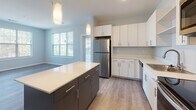 Located at 3 Gilbert in Merrimack, New Hampshire, this single-floor residence offers a thoughtfully designed layout with modern finishes and a cohesive, neutral color palette throughout. The kitchen serves as a standout feature, showcasing a central island with a two-tone design — dark cabinetry below paired with a crisp white countertop — contrasted by bright white upper cabinets lining the walls. Stainless steel appliances, including a refrigerator, dishwasher, electric range, and microwave, provide a polished, functional workspace, while stylish pendant lighting adds a contemporary touch. Natural light filters in through multiple windows, complementing the warm wood-look plank flooring that runs through the main living areas. The bedrooms are carpeted in a neutral tone, each featuring windows that bring in outdoor views and ample natural light, creating comfortable and versatile spaces. Two bathrooms serve the residence, each finished with wood-look tile flooring and modern vanities with clean-lined cabinetry. One bathroom includes a soaking tub, while the other features a walk-in shower. The overall ambiance is clean, contemporary, and move-in ready, with consistent design choices that lend a cohesive feel across every room.