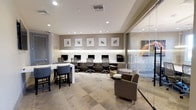 This single-floor business center offers a thoughtfully designed workspace that balances functionality with contemporary style. The room features a row of dedicated computer workstations along a sleek, built-in dark desk running the length of one wall, each equipped with monitors and ergonomic mesh chairs, illuminated by subtle under-cabinet lighting and a series of recessed ceiling lights that create a bright, focused environment. Above the workstations, a curated collection of framed abstract artwork adds visual interest against the neutral taupe walls. A built-in ledge with textured stone detailing serves as an attractive backdrop, enhancing the refined aesthetic of the space. To one side, a high counter-height table paired with upholstered bar stools provides an alternative work surface ideal for quick tasks or informal collaboration. Comfortable lounge seating with a small round side table offers a more relaxed area for casual meetings or reading. A floor-to-ceiling glass partition separates an adjacent meeting or private office space, maintaining an open and airy feel while providing visual privacy. The patterned carpet tile flooring ties the various zones together cohesively, contributing to the space's professional yet welcoming ambiance.