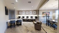 This single-floor business center offers a thoughtfully designed workspace that balances functionality with contemporary style. The room features a row of dedicated computer workstations along a sleek, built-in dark desk running the length of one wall, each equipped with monitors and ergonomic mesh chairs, illuminated by subtle under-cabinet lighting and a series of recessed ceiling lights that create a bright, focused environment. Above the workstations, a curated collection of framed abstract artwork adds visual interest against the neutral taupe walls. A built-in ledge with textured stone detailing serves as an attractive backdrop, enhancing the refined aesthetic of the space. To one side, a high counter-height table paired with upholstered bar stools provides an alternative work surface ideal for quick tasks or informal collaboration. Comfortable lounge seating with a small round side table offers a more relaxed area for casual meetings or reading. A floor-to-ceiling glass partition separates an adjacent meeting or private office space, maintaining an open and airy feel while providing visual privacy. The patterned carpet tile flooring ties the various zones together cohesively, contributing to the space's professional yet welcoming ambiance.