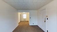 This single-floor residential space offers a simple, functional layout designed for comfortable everyday living. The interior is finished with wall-to-wall carpeting in a warm neutral tone throughout the main living areas, providing a cozy underfoot feel. The ceiling features a distinctive textured tile pattern, adding a layer of visual detail to the otherwise clean, white-walled rooms. The open flow between rooms is framed by a wide doorway, creating a natural sense of continuity and allowing natural light to travel through the space. A front-facing window in the adjoining room brings in daylight, brightening the interior and contributing to a light, airy atmosphere. White painted walls and trim run consistently throughout, lending a clean and cohesive aesthetic. A closet with a paneled white door provides built-in storage. Utility elements such as a baseboard radiator are integrated into the layout, reflecting the practical nature of the space. Overall, this unit presents a straightforward, well-maintained interior that offers a solid foundation for personalization and comfortable residential use.