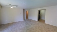 This single-floor space centers around a spacious, open living area finished with wall-to-wall neutral-toned carpeting that creates a clean, understated foundation. The room is bright and airy, with white walls and a standard ceiling equipped with a ceiling fan, providing both light and airflow. The layout is notably open, offering a generous sense of space suitable for a variety of furniture arrangements.

Toward the right side of the living area, an opening leads directly into the kitchen, which features dark cabinetry contrasted against light-colored flooring — likely hardwood or laminate — creating a visual distinction between the two spaces. The kitchen appears functional and well-appointed with dark fixtures and ample cabinet storage visible from the living area.

On the left side of the living room, a doorway opens into a hallway that connects to additional rooms beyond, suggesting a practical and straightforward floor plan. The transitions between spaces are clean and unobstructed, contributing to an efficient flow throughout the unit. Overall, the space presents a simple, well-maintained interior with a neutral palette that offers a versatile canvas for personalization.
