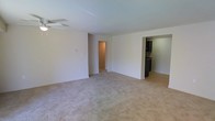 This single-floor space centers around a spacious, open living area finished with wall-to-wall neutral-toned carpeting that creates a clean, understated foundation. The room is bright and airy, with white walls and a standard ceiling equipped with a ceiling fan, providing both light and airflow. The layout is notably open, offering a generous sense of space suitable for a variety of furniture arrangements.

Toward the right side of the living area, an opening leads directly into the kitchen, which features dark cabinetry contrasted against light-colored flooring — likely hardwood or laminate — creating a visual distinction between the two spaces. The kitchen appears functional and well-appointed with dark fixtures and ample cabinet storage visible from the living area.

On the left side of the living room, a doorway opens into a hallway that connects to additional rooms beyond, suggesting a practical and straightforward floor plan. The transitions between spaces are clean and unobstructed, contributing to an efficient flow throughout the unit. Overall, the space presents a simple, well-maintained interior with a neutral palette that offers a versatile canvas for personalization.