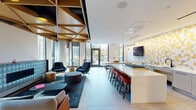 Located at 845 W Madison St in Chicago's vibrant West Loop neighborhood, this single-floor amenity lounge is a thoughtfully designed communal space that blends modern aesthetics with a welcoming atmosphere. The space is anchored by a striking geometric wood-paneled ceiling that adds warmth and visual depth throughout, complemented by large floor-to-ceiling windows that flood the interior with natural light and offer views of the surrounding cityscape and landscaped greenery.

The lounge area features multiple distinct seating arrangements, including plush modular sofas and accent chairs in deep blues and neutral tones, arranged around a linear fireplace clad in a decorative teal geometric tile surround — a true focal point of the space. A dedicated TV lounge area offers a large wall-mounted screen paired with comfortable sectional seating.

A full-length kitchen bar with a white countertop, professional-grade gas range, and bold red barstools serves as the social hub of the space, accented by a gold-and-white patterned feature wall. Adjacent to the kitchen bar is a dining area with a solid wood communal table and teal chairs, ideal for casual gatherings. A shuffleboard table and glass-partitioned rooms add recreational and functional versatility to the overall layout.