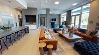 This contemporary single-floor amenity space is designed with an open, flowing layout that seamlessly connects its key functional zones. Floor-to-ceiling glass walls and oversized entry doors allow abundant natural light to pour throughout, creating a bright and welcoming atmosphere complemented by neutral tones and modern finishes.

The lounge area serves as the centerpiece of the space, featuring rich cognac leather sofas arranged around a substantial wooden coffee table. A sleek, wall-mounted flat-screen television is positioned above a linear electric fireplace set into a crisp white surround, providing a stylish and cozy focal point. Accent chairs in deep navy blue add a sophisticated contrast to the warm leather seating.

Adjacent to the lounge, the full-scale community kitchen is a standout feature. It is outfitted with high-gloss white cabinetry, a glass tile backsplash, stainless steel appliances including a professional-grade double wall oven and an oversized refrigerator, and a gas cooktop with a ventilation hood. A large island with a dark countertop and gold-accented bar stools provides generous counter seating, making the space ideal for gatherings and events.

Light wood-toned flooring runs through the lounge area, while large-format tile anchors the kitchen. The overall design balances modern sophistication with comfort, making this an exceptionally functional and visually appealing shared amenity space.