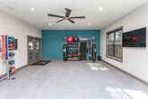Located in Crest Village in Flowery Branch, GA, this single-floor fitness center offers a comprehensive workout environment designed to support a variety of exercise routines. The space is divided into distinct functional zones, each thoughtfully outfitted with quality equipment. One area features a full rack of dumbbells alongside stationary cycling bikes, bathed in natural light streaming through large, framed windows that overlook the surrounding greenery. Large wall-mounted mirrors amplify the sense of openness and allow for form-checking during workouts. A bold teal accent wall anchors a dedicated functional training zone, complete with a multi-use rig holding punching bags, kettlebells, battle ropes, and storage for medicine balls and fitness accessories. Additional equipment includes stability balls in multiple sizes, resistance bands, and a medicine ball rack. Overhead, a striking large-blade ceiling fan provides airflow throughout the space, complemented by recessed lighting for consistent illumination. The flooring throughout is a neutral, textured carpet tile suited for both light cardio and strength training. Glass-paneled doors connect the workout zones, enhancing the flow between areas. This fitness center is a well-rounded facility offering everything needed for a complete, full-body workout experience.