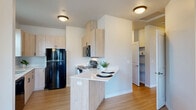 This single-floor residence offers four bedrooms and two bathrooms within a thoughtfully designed layout. Light oak-toned laminate flooring runs continuously throughout, creating a cohesive and clean aesthetic across every room. The kitchen is outfitted with light wood cabinetry, white quartz countertops, and black appliances including a refrigerator, electric range with over-the-range microwave, and a peninsula with bar seating — ideal for casual dining or entertaining. A dedicated dining and living area sits adjacent to the kitchen, featuring vaulted ceilings, a stylish chandelier, and multiple windows that invite generous natural light into the space. The four bedrooms are well-proportioned, with the spacious primary bedroom offering vaulted ceilings. The secondary bedrooms are bright and functional, each with windows overlooking the surrounding neighborhood. Both bathrooms are finished with light wood vanities, white countertops, large mirrors, and tub-shower combinations. A built-in shelving nook adds convenient storage near the hallway. The overall design palette — neutral walls, warm wood tones, and modern fixtures — creates a clean, contemporary ambiance suitable for a variety of living arrangements.