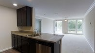 This single-floor residence offers a thoughtfully designed open-concept layout that seamlessly connects the kitchen and living areas, creating a spacious and airy feel throughout. The kitchen is a standout feature, showcasing rich dark cabinetry that provides ample storage, complemented by granite-style countertops and a central peninsula with a stainless steel sink — ideal for both meal preparation and casual dining. A stainless steel dishwasher is integrated into the cabinetry, contributing to a sleek, cohesive aesthetic. Crown molding runs along the ceiling, adding a refined architectural detail that elevates the overall design. The living area is bathed in natural light, courtesy of large windows that frame views of lush greenery outside. A sliding glass door opens directly onto a private balcony, seamlessly blending indoor and outdoor living. The living space features plush wall-to-wall carpeting, offering comfort underfoot, while neutral wall tones throughout create a clean, versatile backdrop. Recessed lighting and a pendant light fixture further enhance the welcoming ambiance. An additional room visible from the main living area suggests flexible use of space, whether as a bedroom, office, or den. Overall, the residence balances modern finishes with functional design.