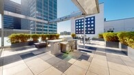 Located atop a building along the iconic Wilshire Boulevard corridor in Los Angeles, this rooftop terrace offers a sophisticated outdoor amenity space with commanding views of the surrounding Koreatown and Mid-Wilshire skyline. The deck is finished with large-format porcelain tile laid in a clean grid pattern, lending a polished, contemporary aesthetic throughout.

The terrace is thoughtfully organized into multiple functional zones. A dedicated outdoor kitchen area features a stainless steel BBQ grill and prep counter, ideal for entertaining. Adjacent to it, multiple dining tables with chairs are arranged beneath sleek pergola-style shade structures with metal canopies, providing comfortable al fresco seating options. A large-scale oversized Connect Four game adds a playful, social element to the space.

Lush greenery is integrated throughout via generous raised planters filled with bamboo and ornamental shrubs, creating natural privacy screens and softening the urban hardscape. The entry corridor is flanked by tall bamboo planters, offering a welcoming, garden-like transition onto the deck.

The terrace benefits from abundant Southern California sunshine, with open sightlines offering panoramic views of neighboring high-rise office towers and the broader Los Angeles cityscape. The combination of thoughtful landscaping, dining amenities, and recreational features makes this rooftop an exceptional outdoor gathering destination.
