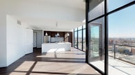 Located on the 17th floor at 300 14th Street in San Diego, this single-level unit offers a striking open-concept layout defined by sleek, contemporary design. The most commanding feature is the expansive floor-to-ceiling glass curtain wall that spans the living area, flooding the interior with abundant natural light and framing sweeping panoramic views of the San Diego cityscape.

The open-plan living and kitchen area flows seamlessly, anchored by warm hardwood-style flooring that complements the clean white walls and high ceilings. The kitchen is thoughtfully appointed with rich, dark wood cabinetry, light grey subway tile backsplash, stainless steel appliances including a refrigerator and microwave, and a generous white waterfall-edge island that provides both prep space and casual seating. Pendant lighting hangs above the island, adding refined detail to the space.

The overall ambiance is modern and airy, with a neutral palette that allows the dramatic city views to serve as the focal point. Large black-framed sliding glass doors provide direct access to an outdoor balcony, further connecting the interior to the surrounding urban environment.