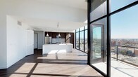 Located on the 17th floor at 300 14th Street in San Diego, this single-level unit offers a striking open-concept layout defined by sleek, contemporary design. The most commanding feature is the expansive floor-to-ceiling glass curtain wall that spans the living area, flooding the interior with abundant natural light and framing sweeping panoramic views of the San Diego cityscape.

The open-plan living and kitchen area flows seamlessly, anchored by warm hardwood-style flooring that complements the clean white walls and high ceilings. The kitchen is thoughtfully appointed with rich, dark wood cabinetry, light grey subway tile backsplash, stainless steel appliances including a refrigerator and microwave, and a generous white waterfall-edge island that provides both prep space and casual seating. Pendant lighting hangs above the island, adding refined detail to the space.

The overall ambiance is modern and airy, with a neutral palette that allows the dramatic city views to serve as the focal point. Large black-framed sliding glass doors provide direct access to an outdoor balcony, further connecting the interior to the surrounding urban environment.